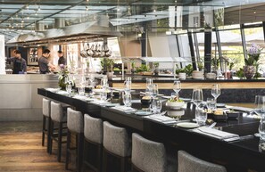 A modern restaurant with a long bar area, neatly set tables, and a kitchen in the background. - Hyatt Regency Mainz (Mainz)