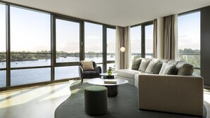 A spacious living room with large windows, a sofa, a chair, a coffee table, and a view of a river. - Hyatt Regency Mainz (Mainz)