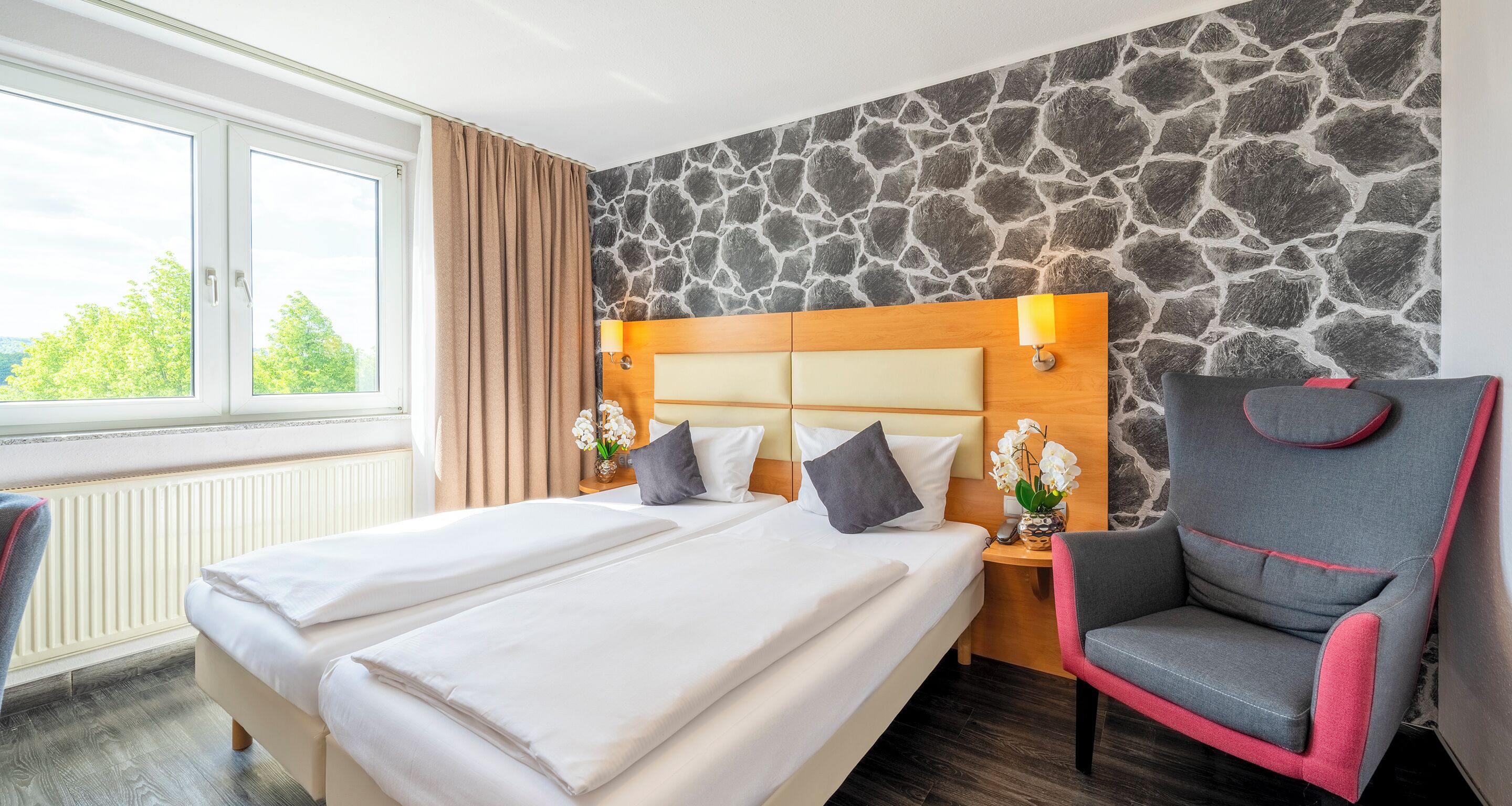 A hotel room with a large bed, a chair, a stone wall, and a window with curtains.