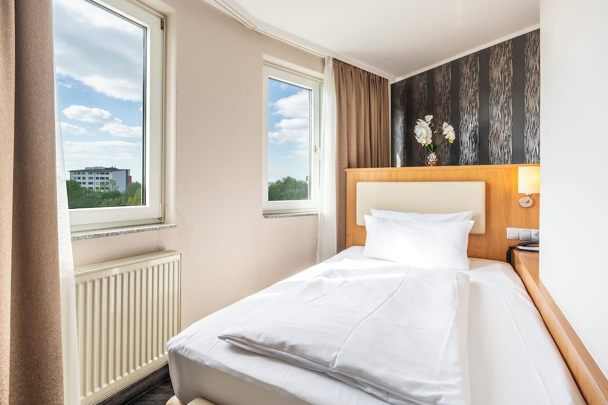 A hotel room with a bed, a nightstand, a lamp, a window, and a view of the outside.