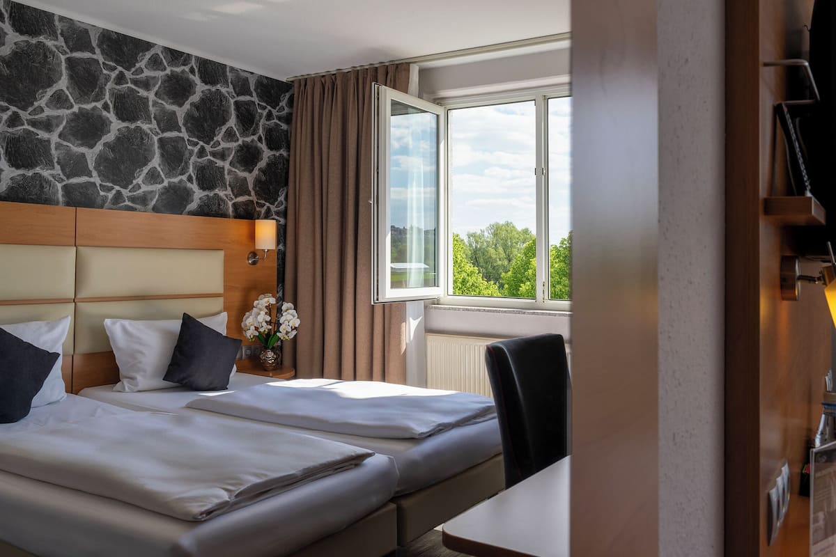 A hotel room with two beds, a stone wall, a window with a view, and a desk with a lamp and a bottle.