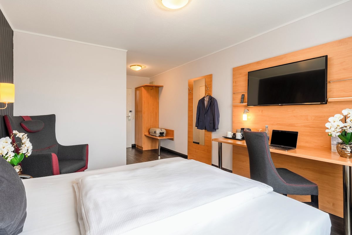 A hotel room with a bed, a desk, a chair, a TV, and a small table with a vase of flowers.