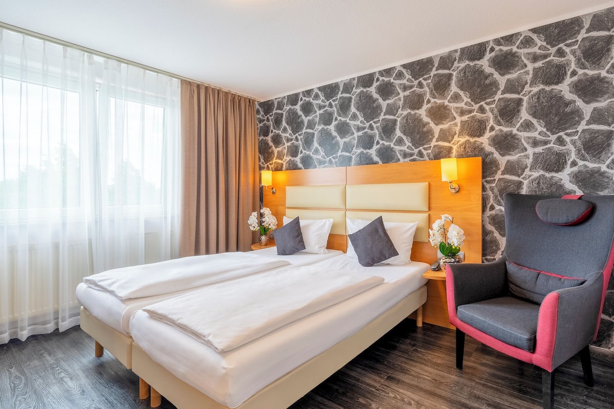 A hotel room with a large bed, a chair, a nightstand, and a stone wall.