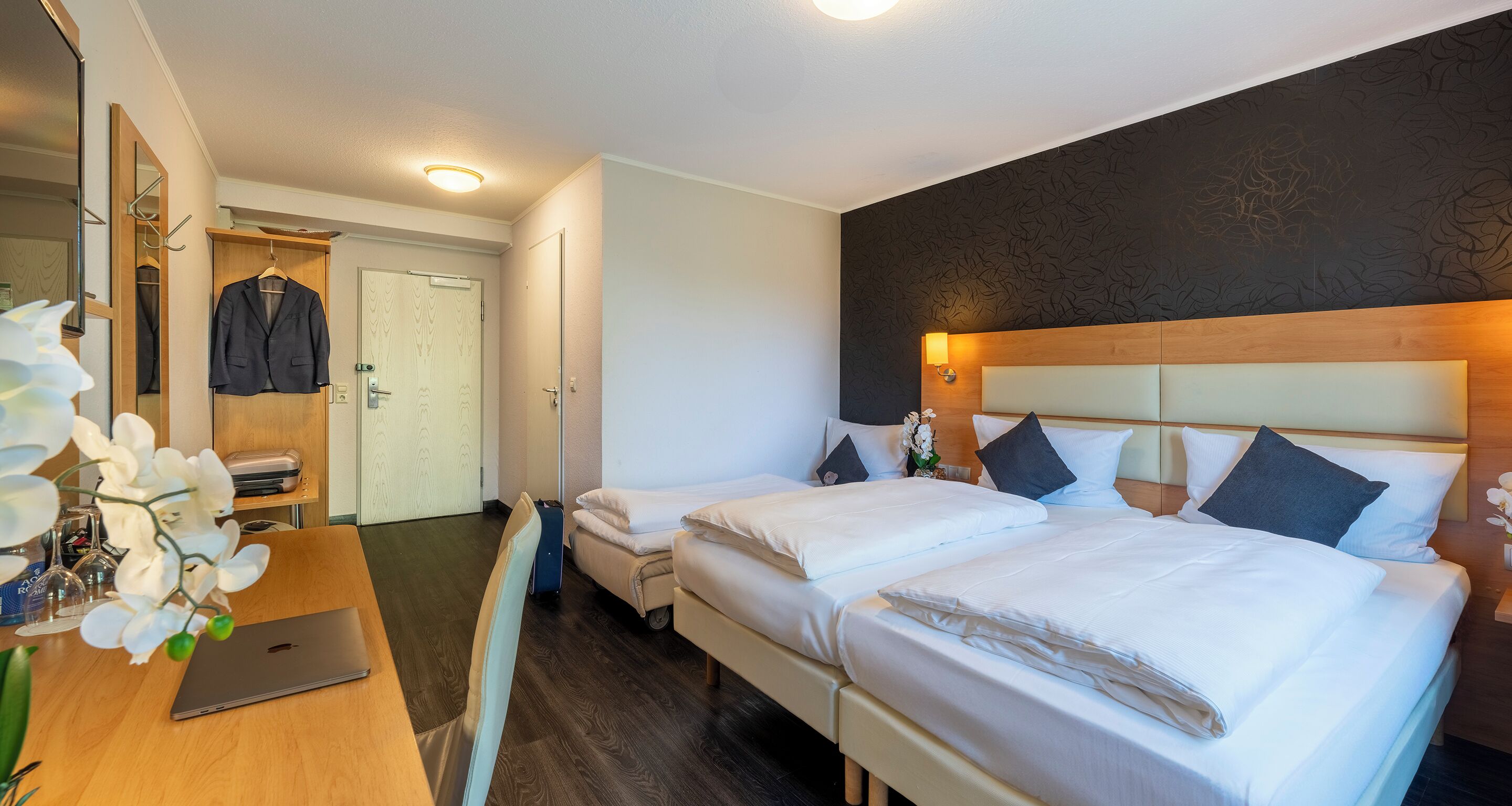 A hotel room with a double bed, a single bed, a desk with a laptop, a mirror, and a wardrobe.