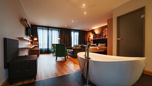 A modern hotel room with a large bathtub, a sofa, a green armchair, and a TV on a shelf. - Der LIPPISCHE HOF (Bad Salzuflen)