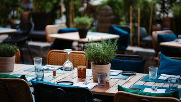 An outdoor dining area with tables set for two, featuring potted plants and clear glassware.
