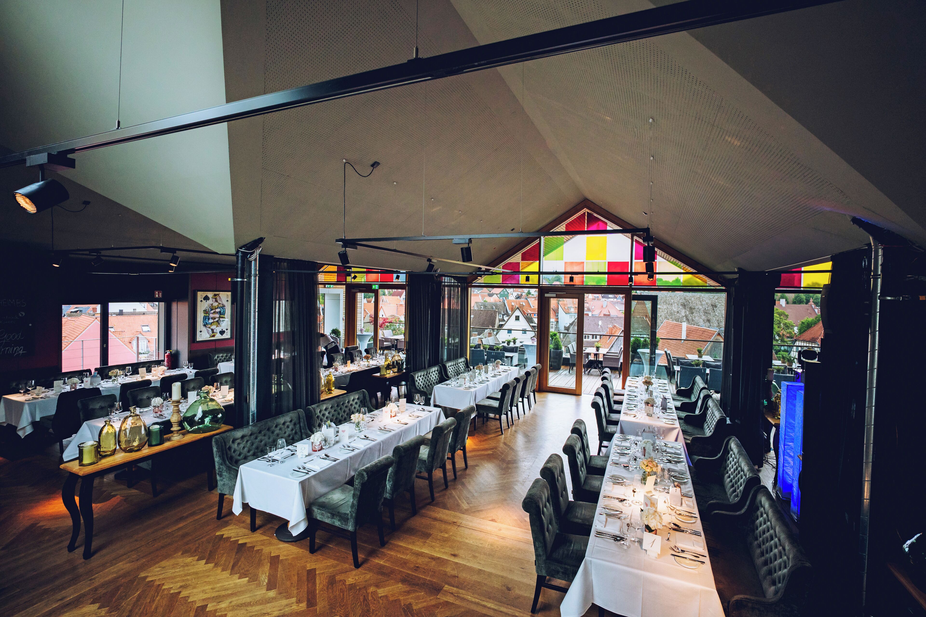 A restaurant with long dining tables, chairs, and a view of buildings through large windows.