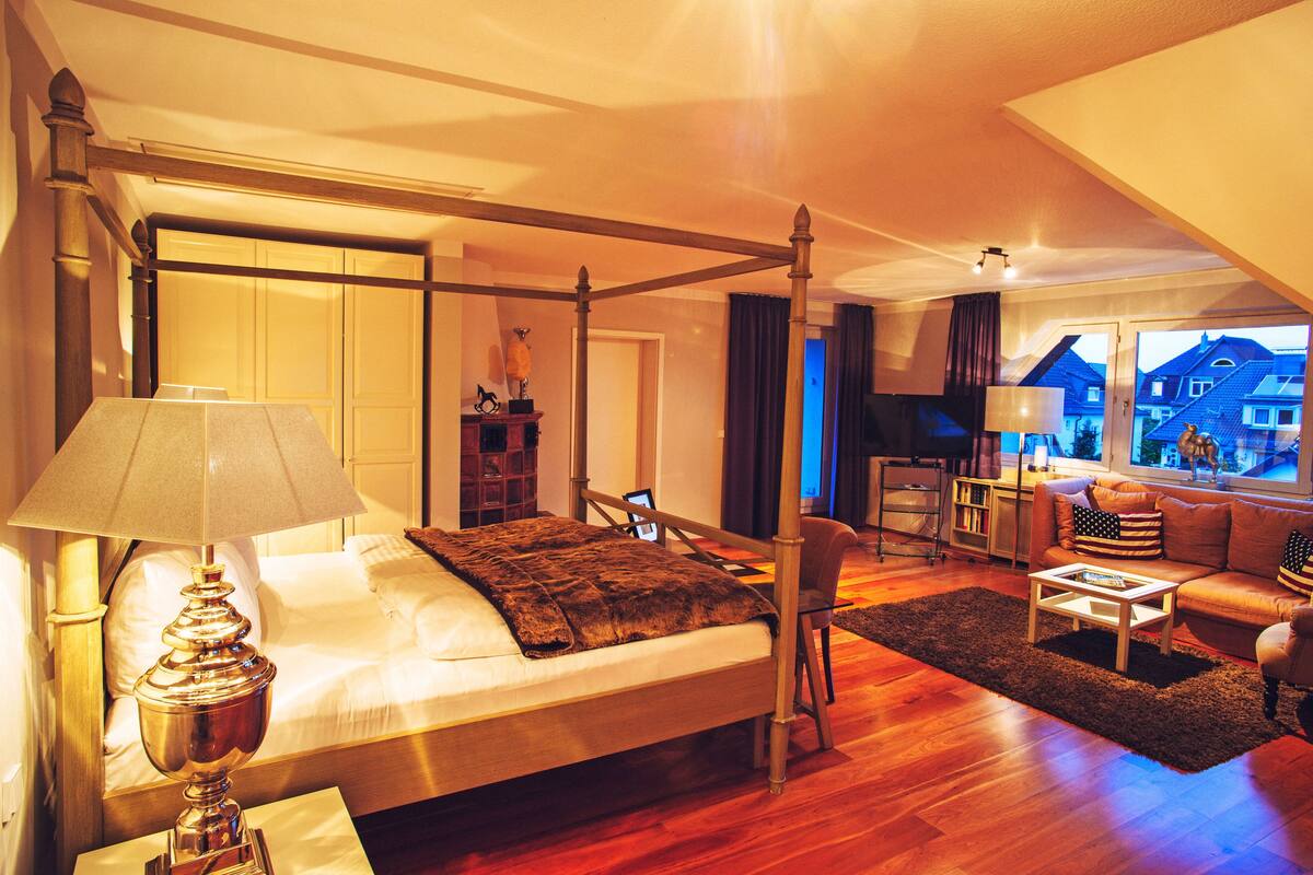 A spacious bedroom with a four-poster bed, a sitting area with a sofa and armchairs, and a view of houses through large windows.