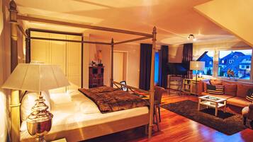 A spacious bedroom with a four-poster bed, a sitting area with a sofa and armchairs, and a view of houses through large windows.
