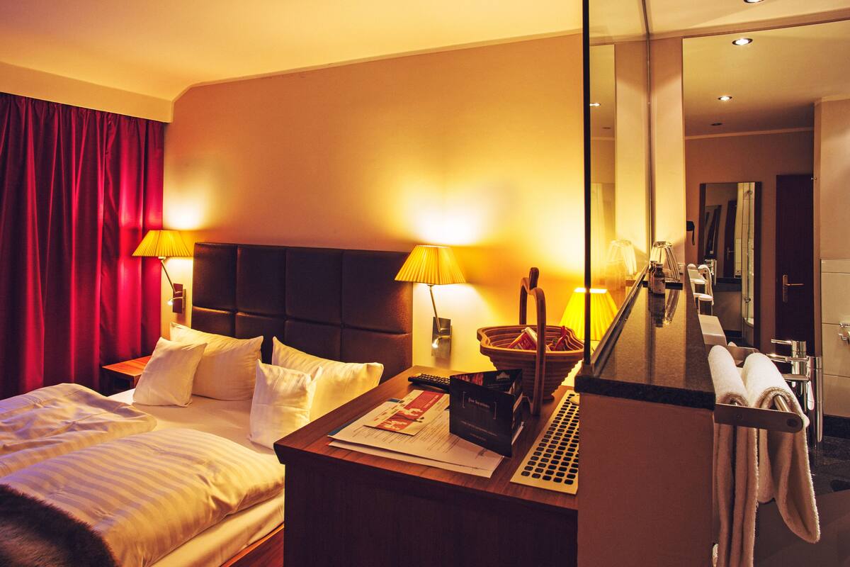 A hotel room with a bed, two bedside lamps, a desk with a lamp, a mirror, and a bathroom visible through an open door.