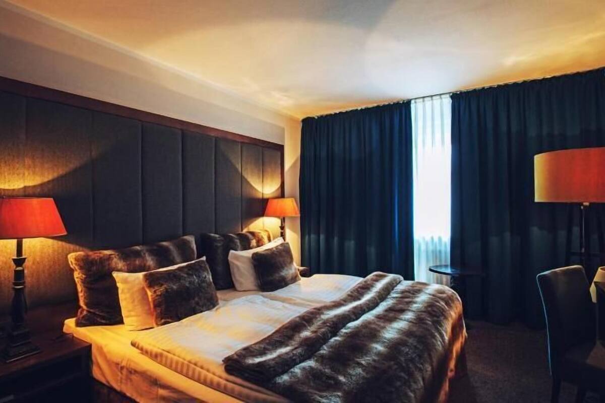A hotel room with a bed, two bedside lamps, a chair, and a window with curtains.