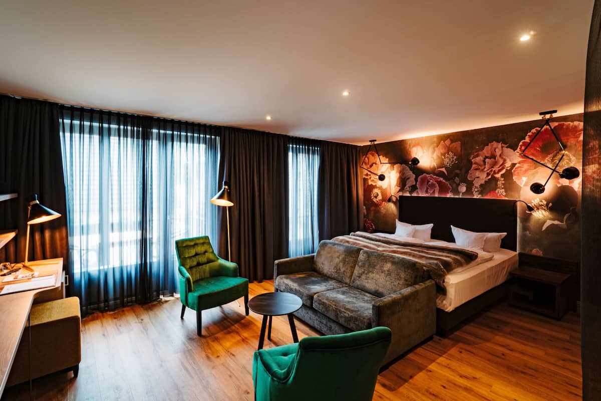 A modern hotel room with a large bed, a green armchair, a sofa, a desk with a lamp, and a large window with curtains.