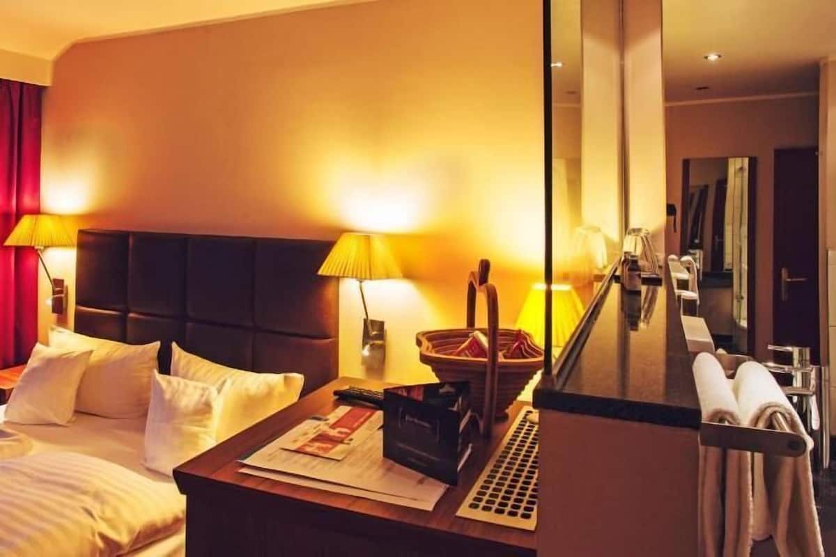 A hotel room with a bed, bedside lamps, a nightstand with a lamp, a mirror, and a towel rack.