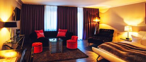 A hotel room with a sofa, armchair, and a bed. There is a small table and a lamp.