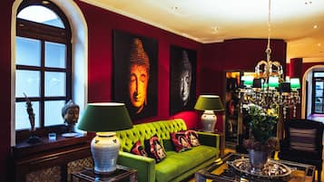 A modern living room with a green sofa, two Buddha paintings, a chandelier, and a window with a decorative Buddha head.
