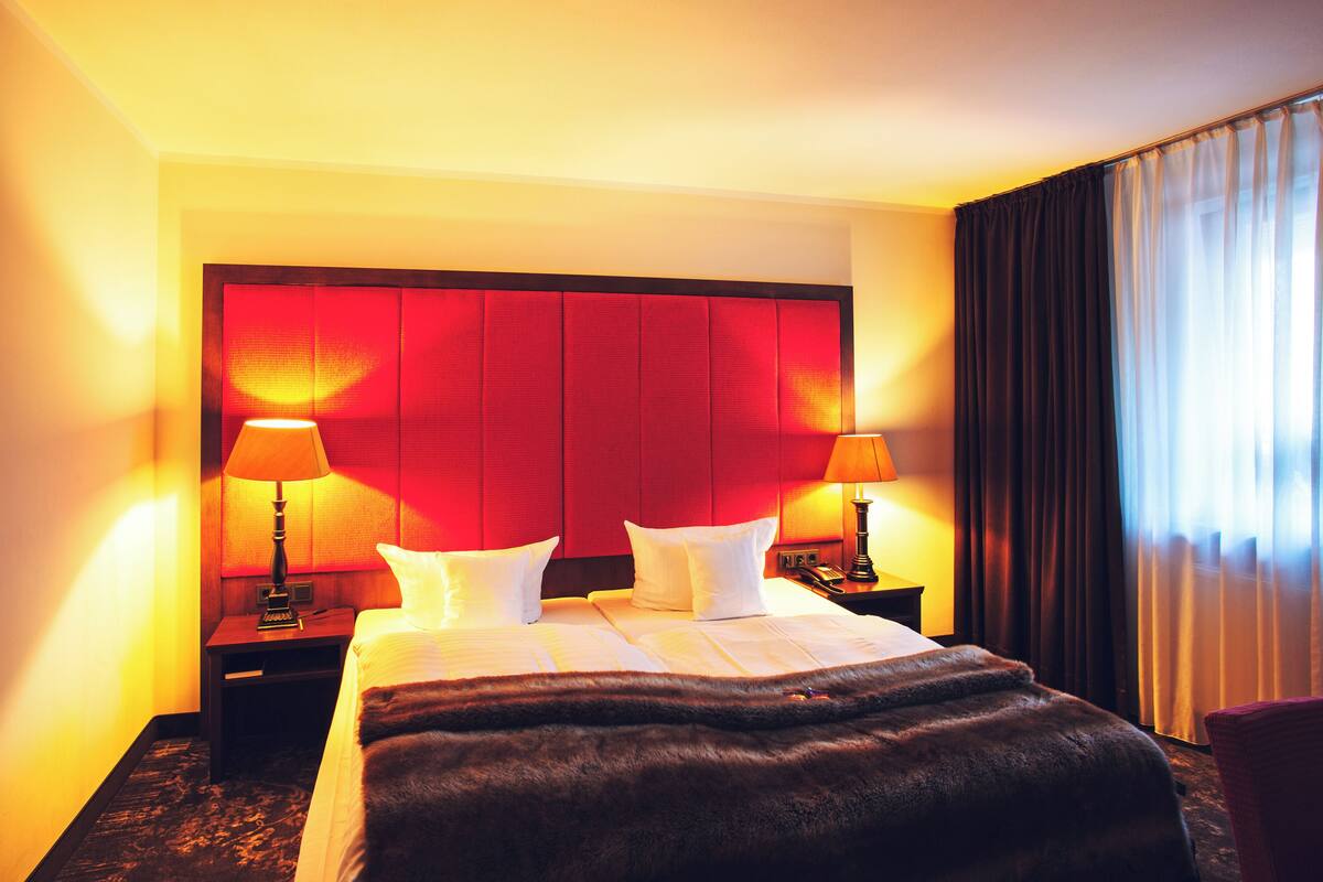 A hotel room with a large bed, two bedside tables with lamps, a red headboard, and a view of the outdoors through a window with curtains.