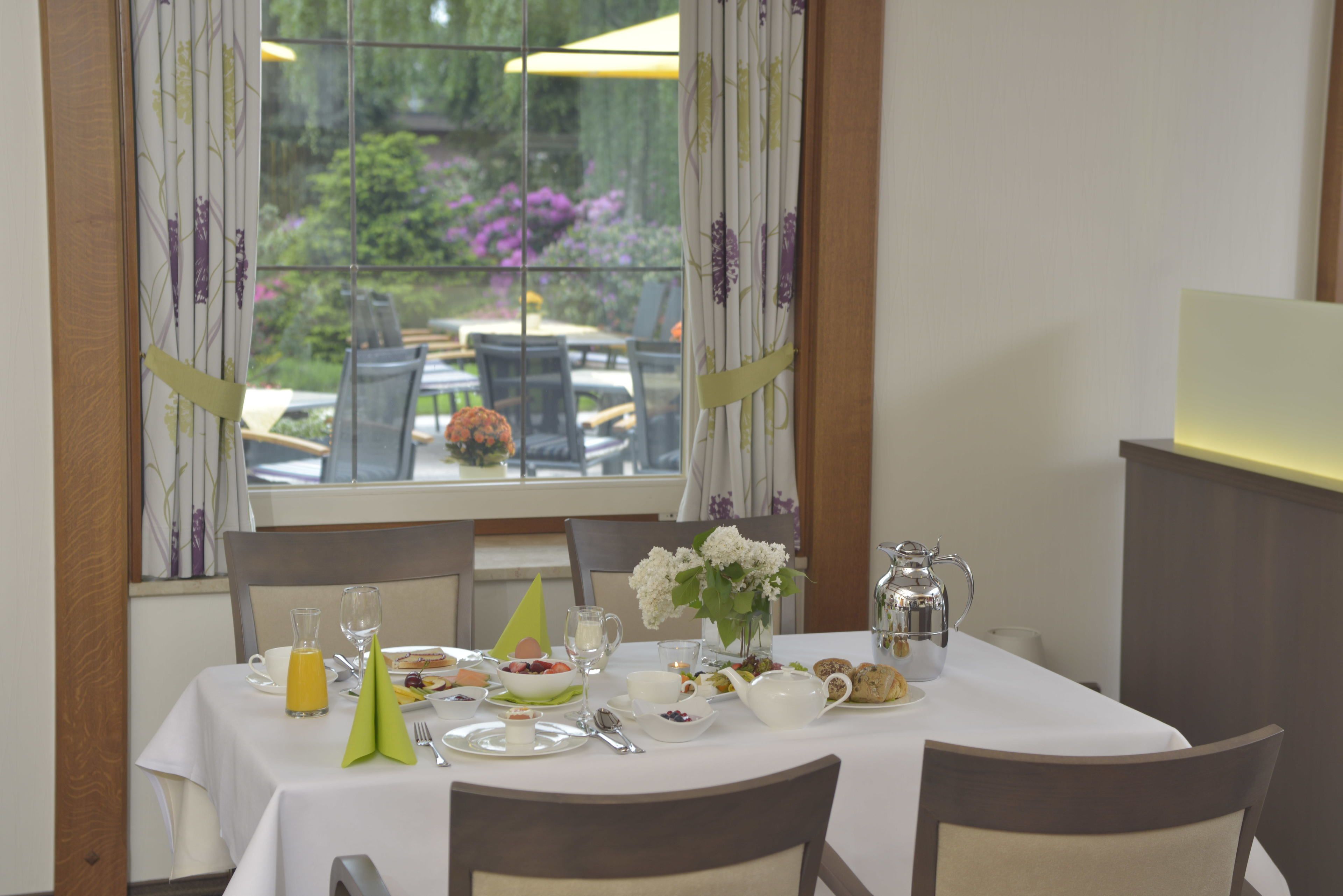 Una mesa de comedor preparada con alimentos para el desayuno, un jarrón de flores y vistas a un jardín exterior a través de una ventana.