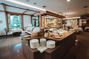 A buffet setup with various food items and utensils, a seating area with tables and chairs, and a view of the outdoors through large windows.
