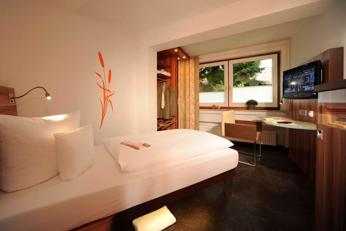 A hotel room with a bed, bedside lamp, a desk with a chair, a television, and a window with curtains.