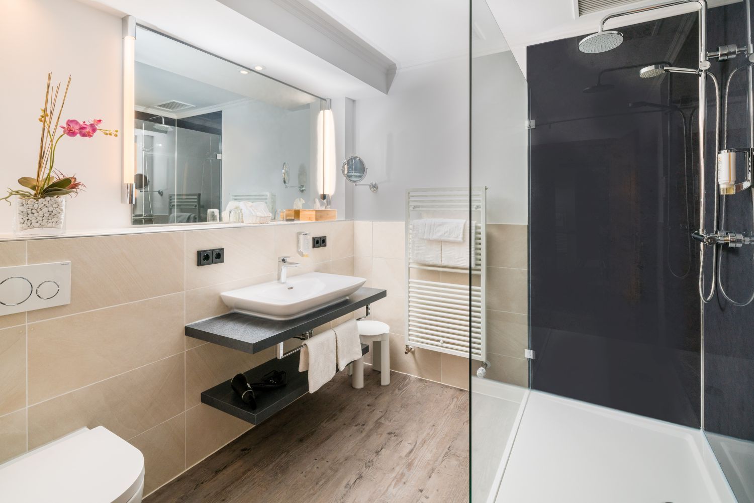 A modern bathroom with a large mirror, a white sink, a glass shower enclosure, and a wooden floor.