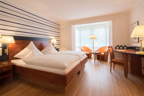 A hotel room with a double bed, two bedside tables with lamps, a desk with a chair, and a window with curtains.