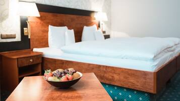 A hotel room with a wooden headboard, a bed with white linens, a nightstand with a lamp, and a table with a bowl of fruit.