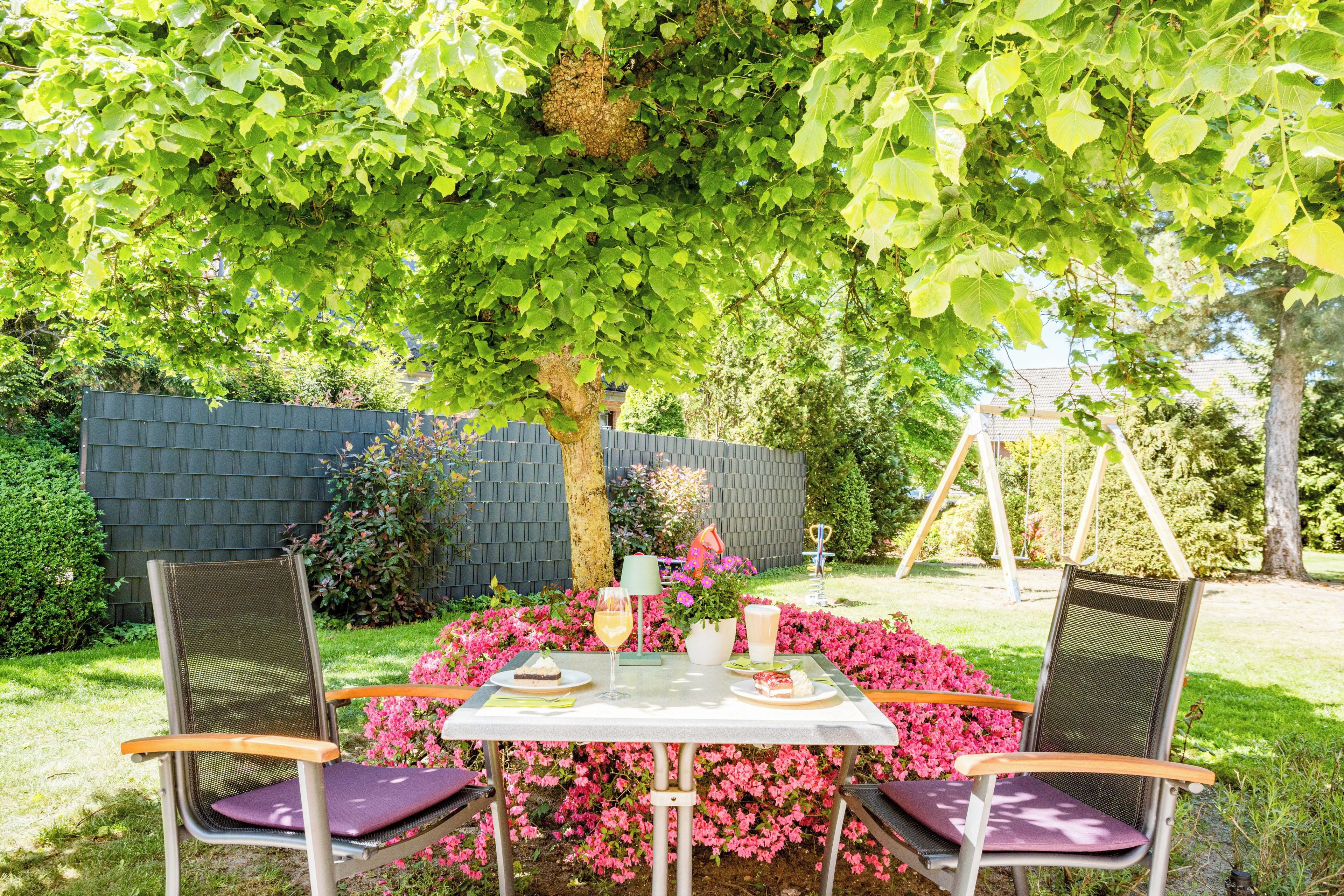 Un espacio de estar al aire libre con una mesa y sillas, rodeado de abundante follaje y plantas con flores.