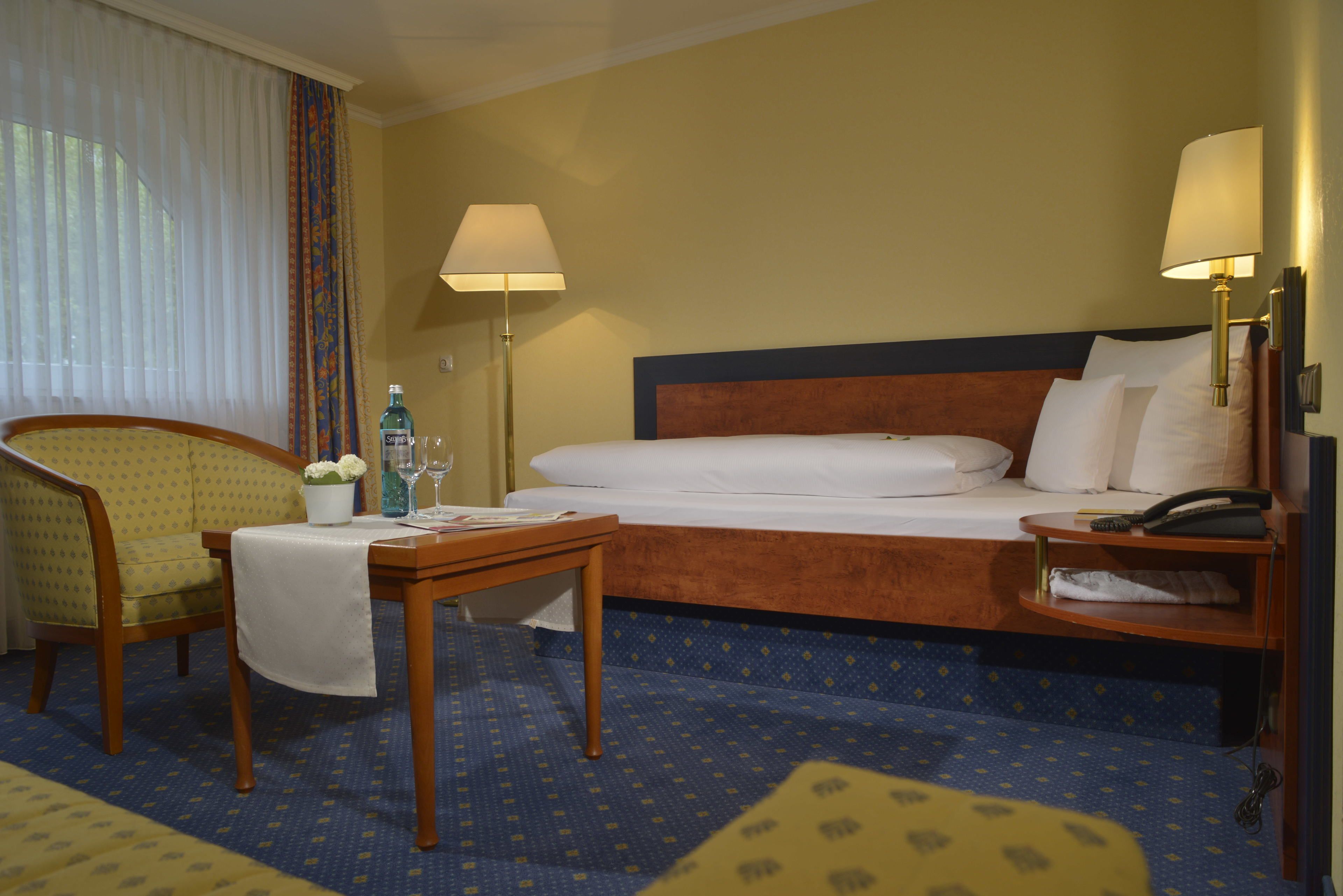 Una habitación de hotel con cama, una silla, una mesa con una botella y vasos, y una lámpara.