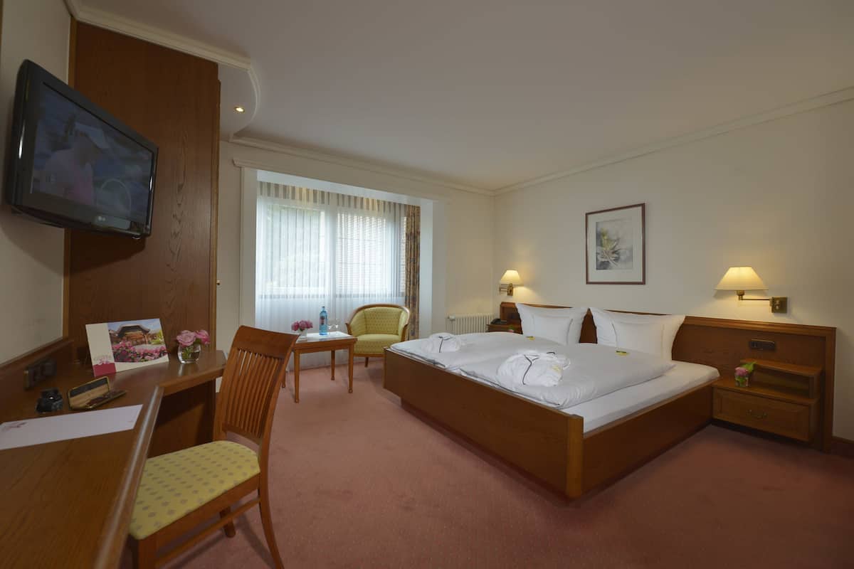 A hotel room with a bed, a desk with a chair, a television, a window with curtains, and a bedside table with a lamp.