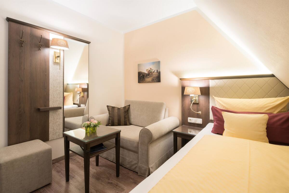 Une chambre d’hôtel avec un lit, un canapé, une petite table sur laquelle se trouve un vase, un miroir et un tableau accroché au mur.