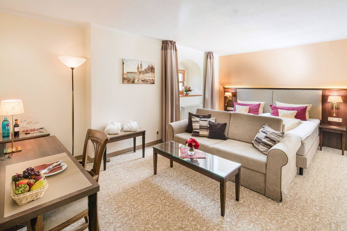Un salon bien éclairé, avec un canapé, une table basse en verre, une table à manger agrémentée d’un panier de fruits et une lampe.