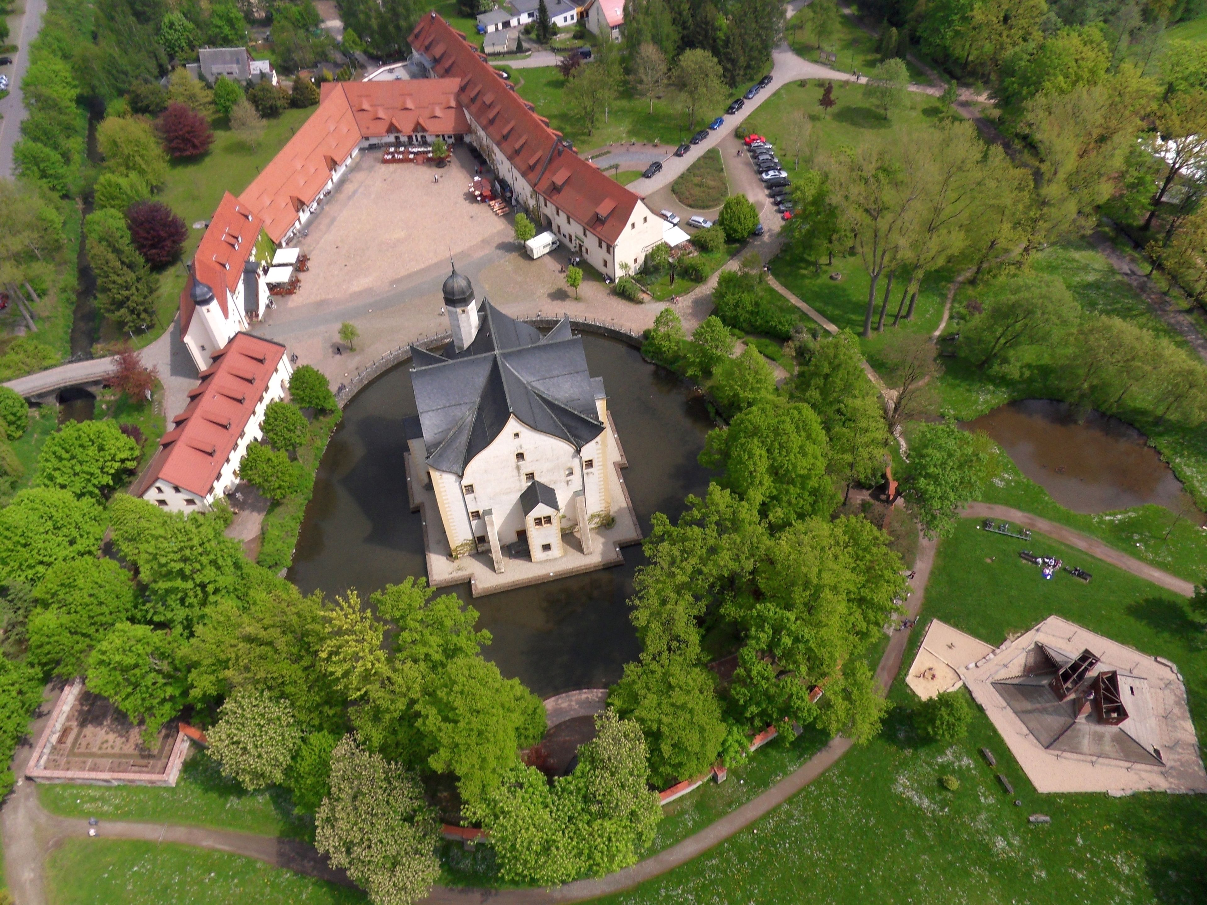 Vista aérea de un complejo de edificios históricos con un patio central, rodeado de abundante vegetación y una pequeña extensión de agua.