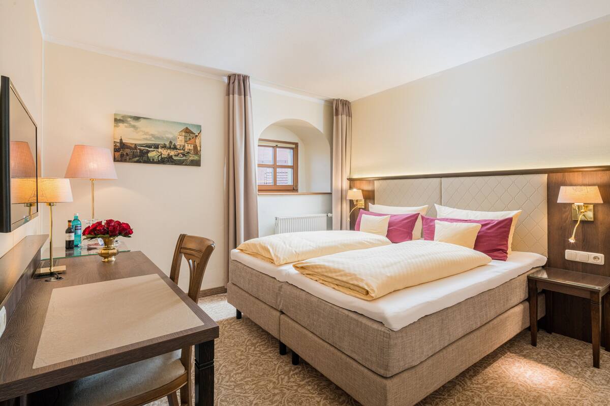 Une chambre d’hôtel avec un lit, un bureau, une chaise, une lampe, un tableau et une fenêtre.