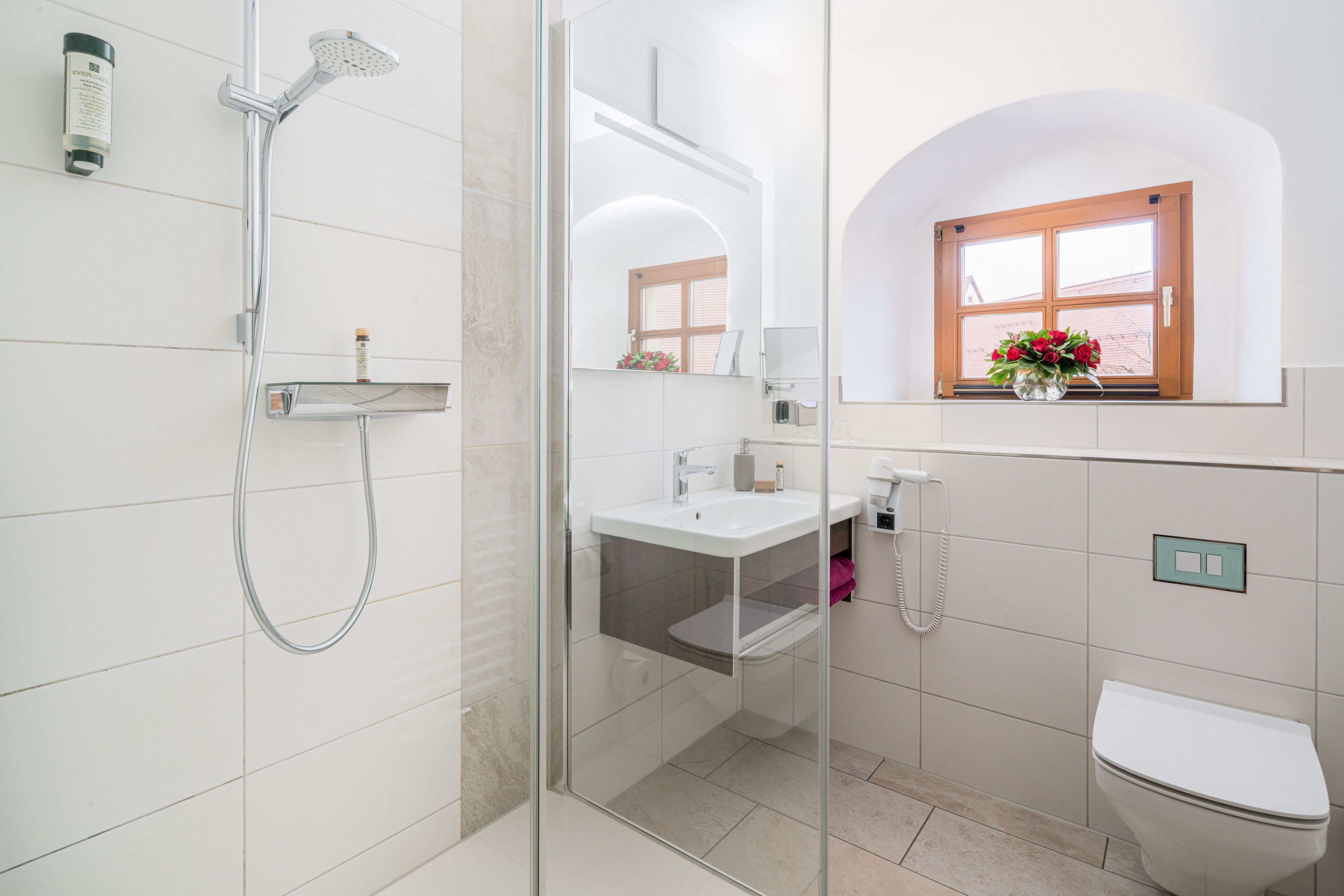 Ein modernes Badezimmer mit einer gläsernen Duschkabine, einem weißen Waschbecken, einer Toilette und einem Fenster mit Blumen.