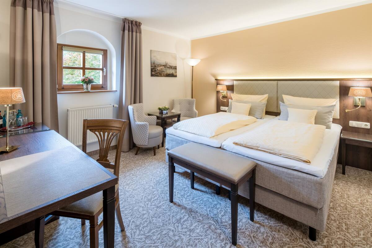 Une chambre d’hôtel avec deux lits, un bureau, une chaise, une petite table et une fenêtre avec des rideaux.