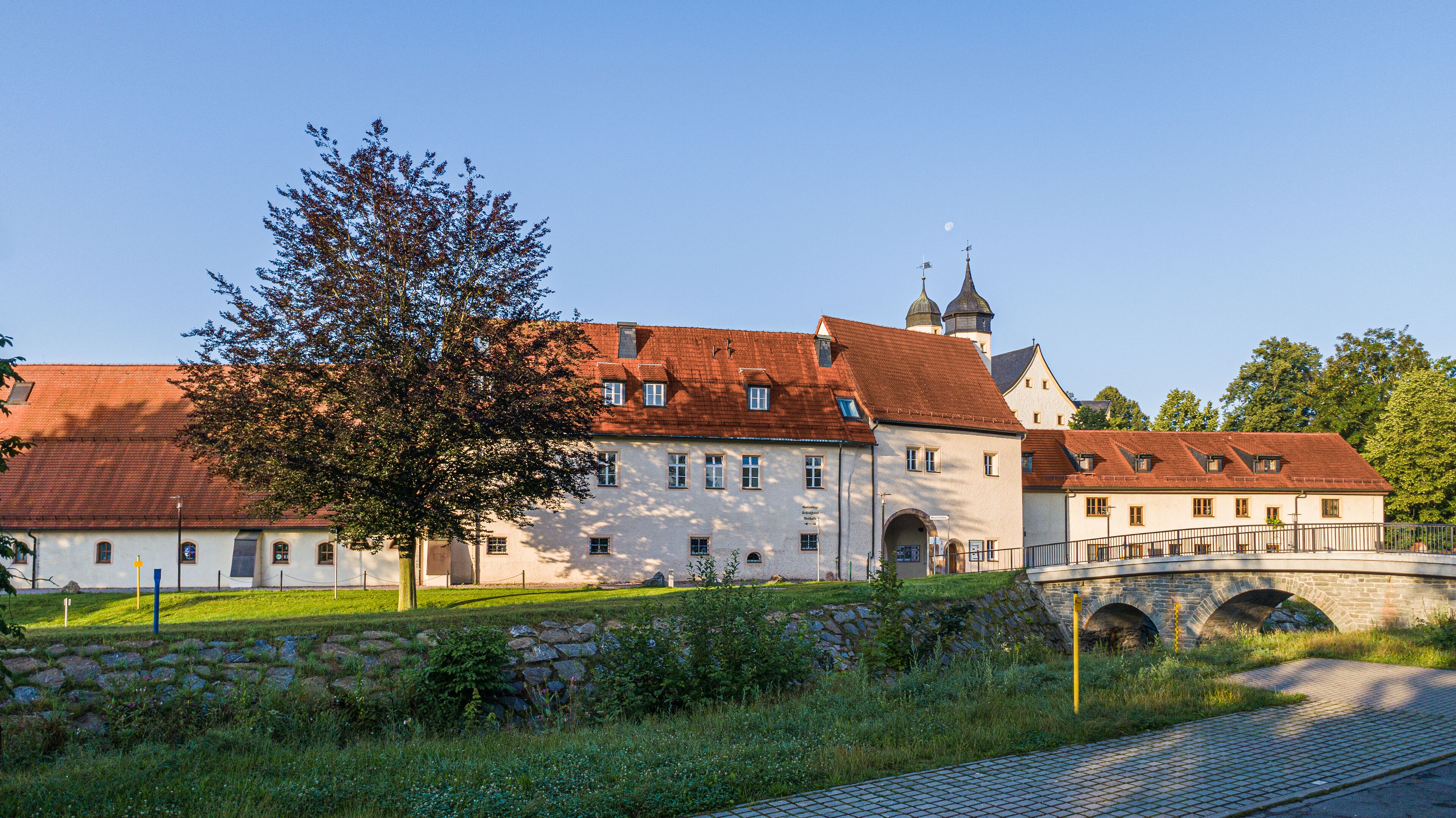 A historic building with a red-tiled roof, a stone bridge, and a large tree in the foreground.