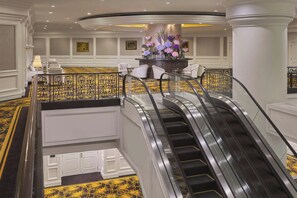 A hotel lobby with an escalator, seating area, reception desk, and decorative floral arrangement.