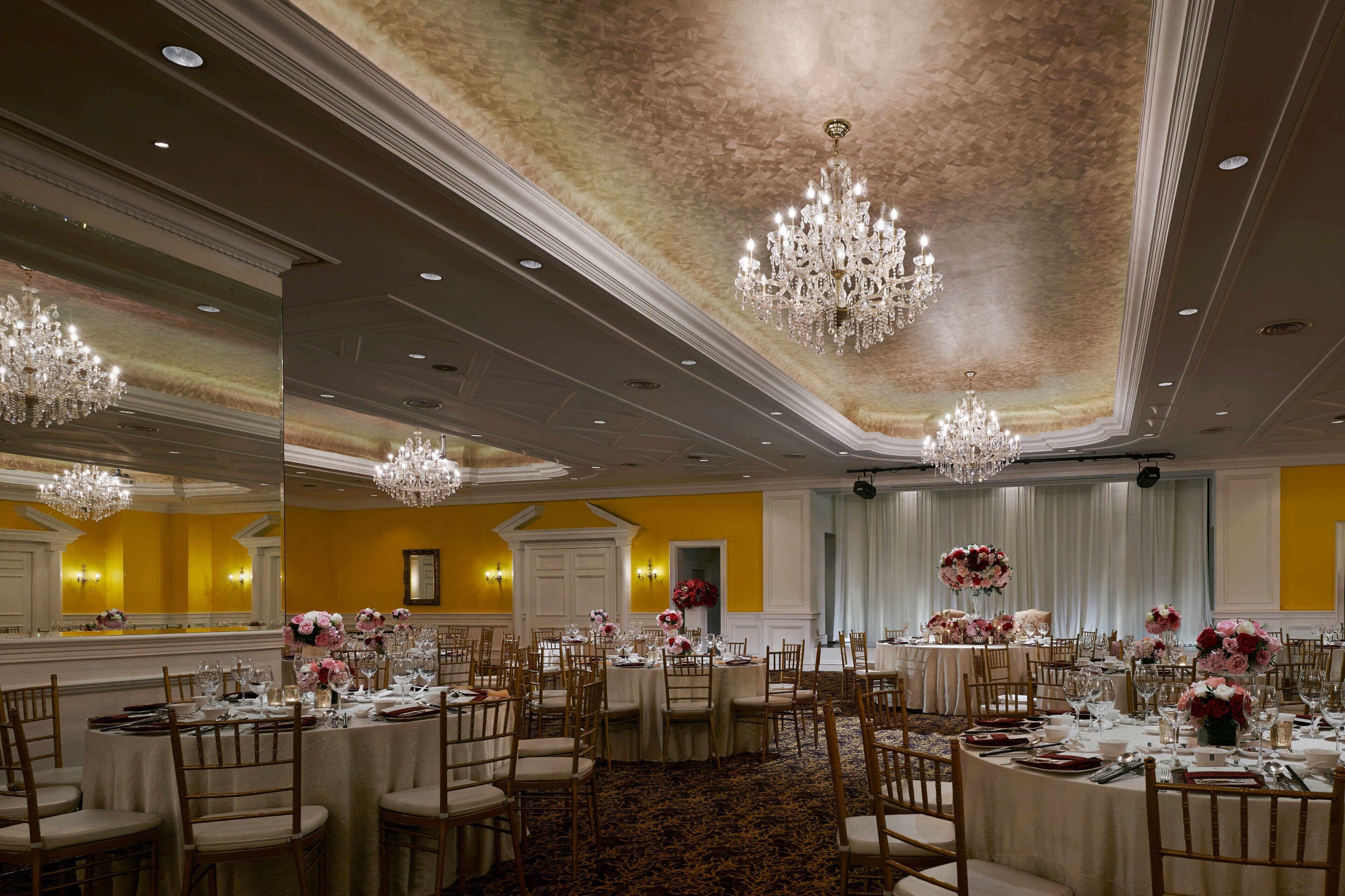 A banquet hall with round tables, chandeliers, and floral centerpieces.