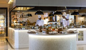 A modern kitchen with chefs preparing food, stainless steel appliances, and a buffet setup.