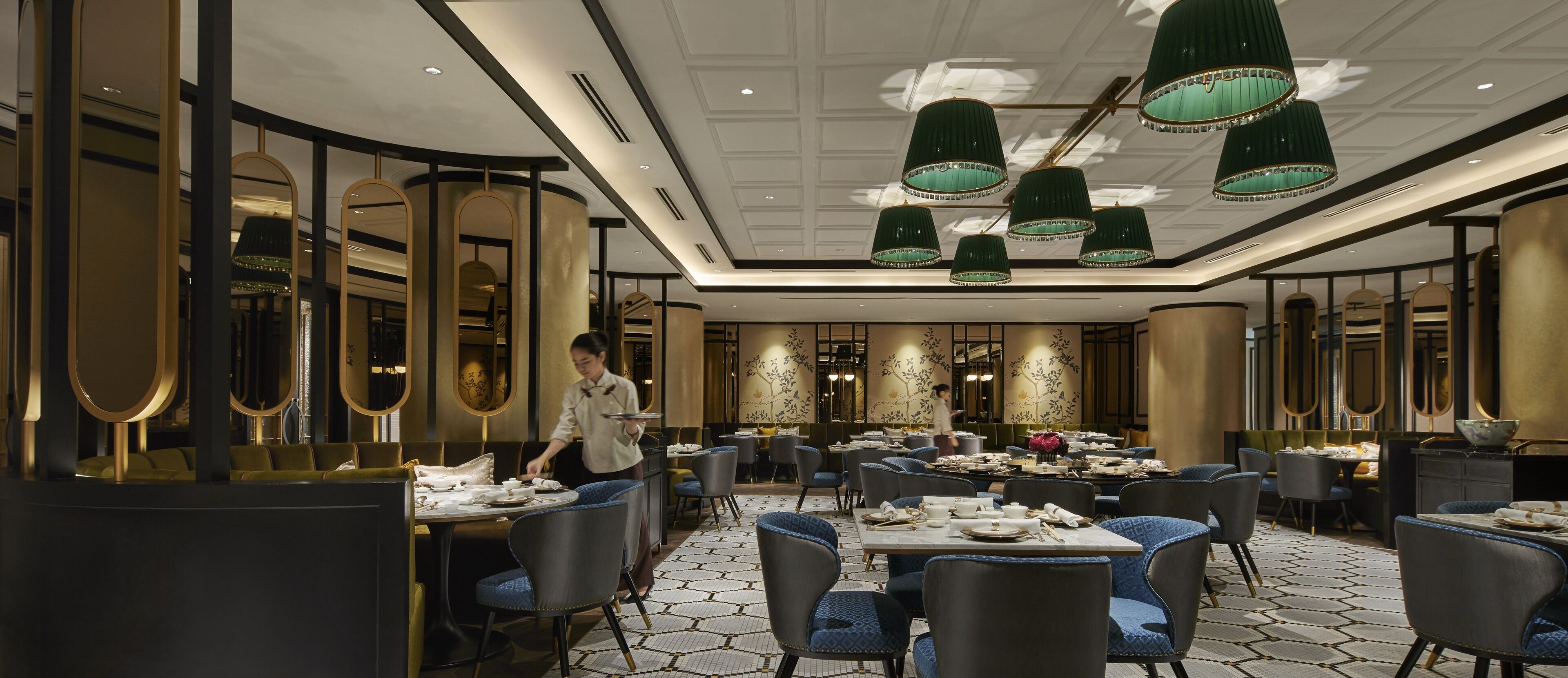 Een restaurant met ronde tafels, blauwe bekleden stoelen en groene hanglampen.