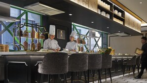 A modern restaurant with a bar area, chefs preparing food, and a person walking by. - The Ritz-Carlton, Kuala Lumpur (Kuala Lumpur)