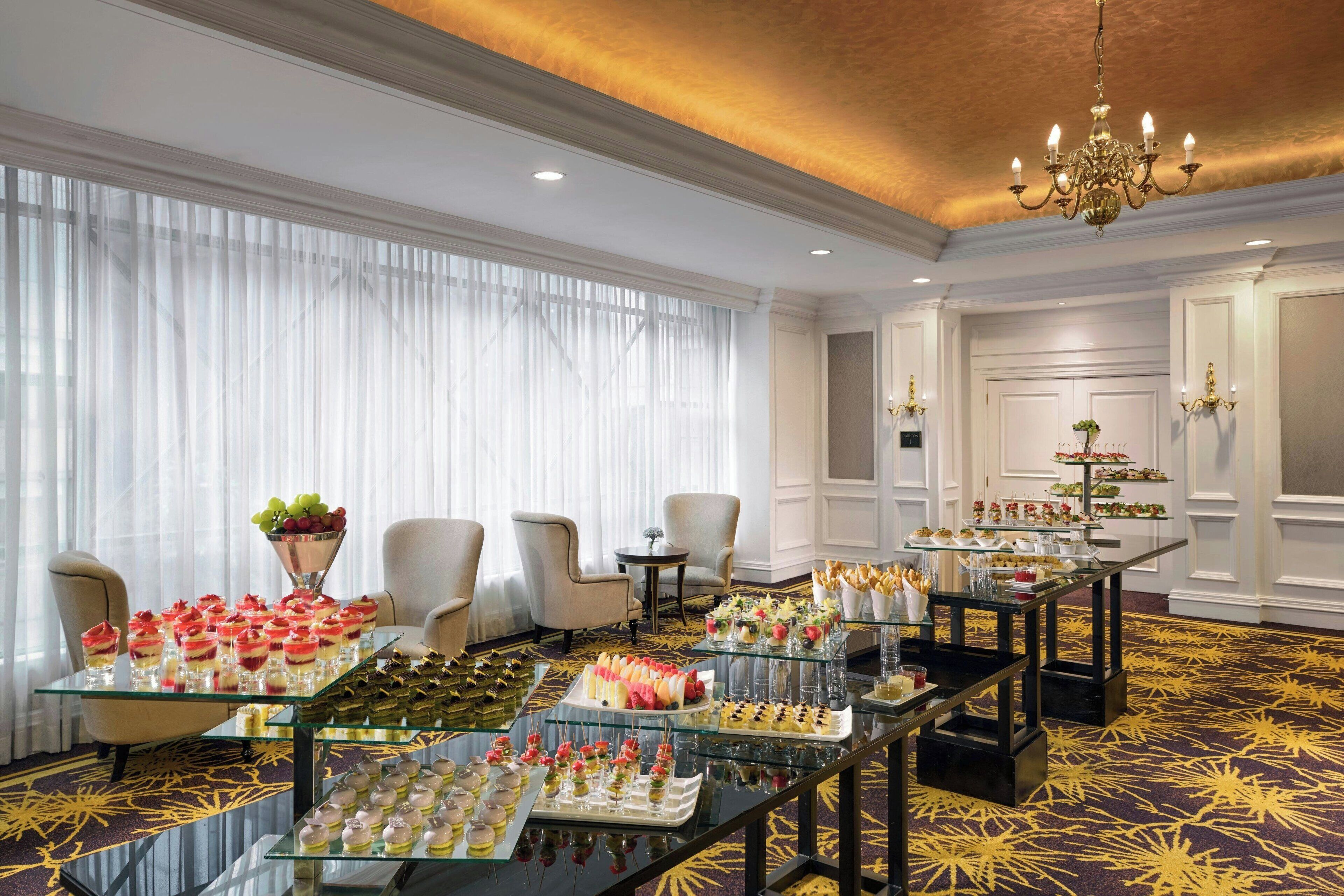 Une chambre d’hôtel luxueuse, dotée d’une grande table à manger dressée avec divers desserts et en-cas, d’élégantes chaises et d’un lustre.