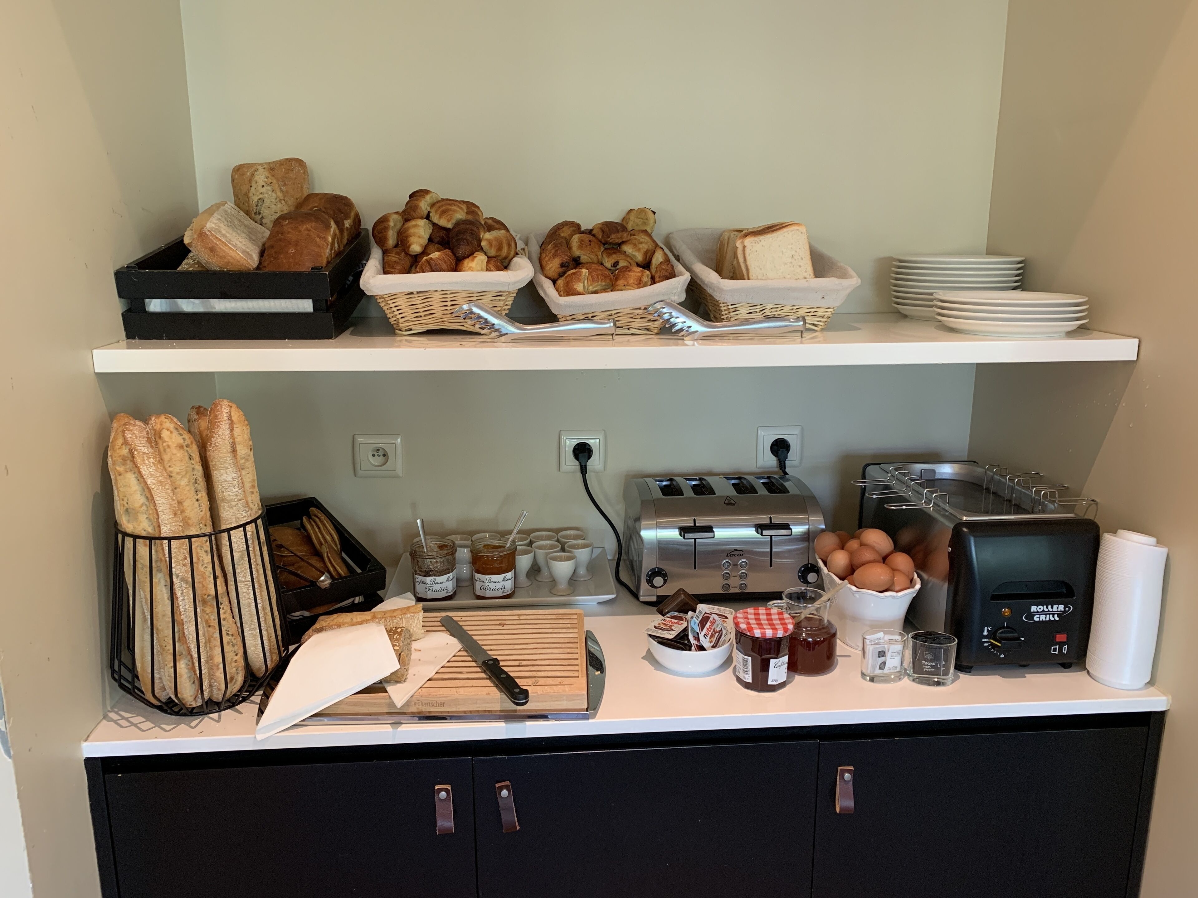 A breakfast buffet setup with bread, croissants, and pastries on the top shelf, and a toaster, eggs, and condiments on the bottom shelf.