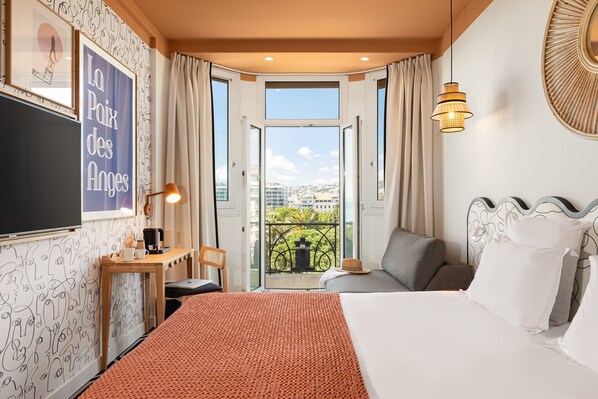 A hotel room with a bed, a desk, a chair, a TV, and a balcony with a view of the city.