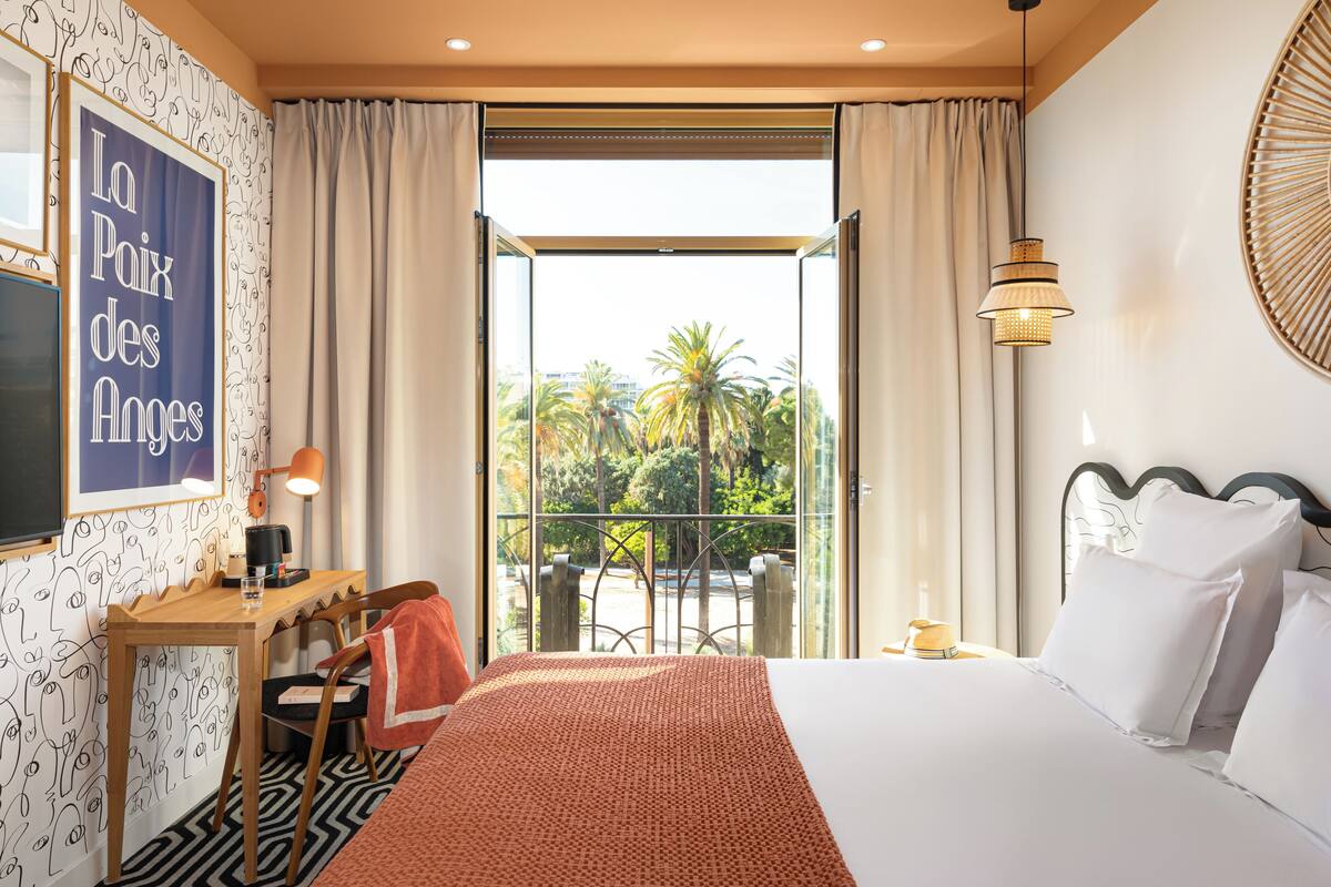 Une chambre d’hôtel avec un lit, un bureau doté d’une lampe, une chaise et une vue sur les palmiers par la fenêtre.
