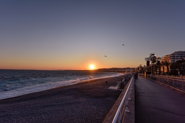A beach with a sunset, a promenade, and buildings in the background.