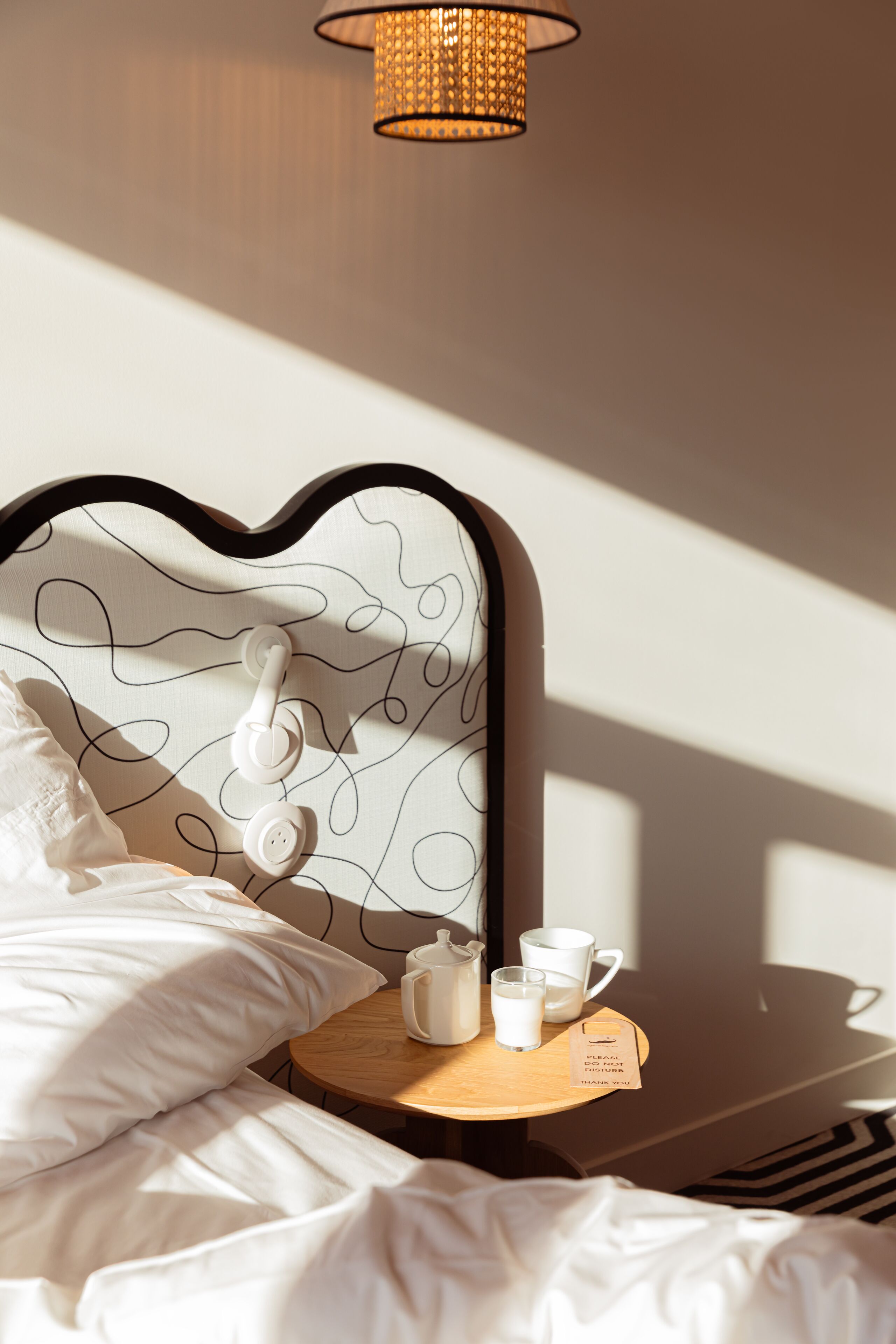 A neatly made bed with white linens, a bedside table with a tray holding a teapot, cups, and a sign.
