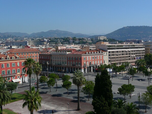 A cityscape with a prominent red building with arched windows and a central plaza with palm trees.