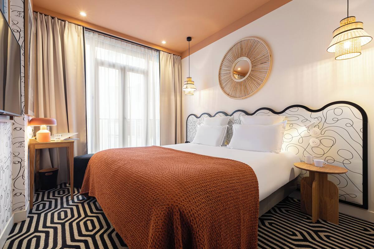 Une chambre d’hôtel avec un lit, une table de chevet avec une lampe, une petite table avec une tasse et une soucoupe, un miroir au mur et un sol à motifs.