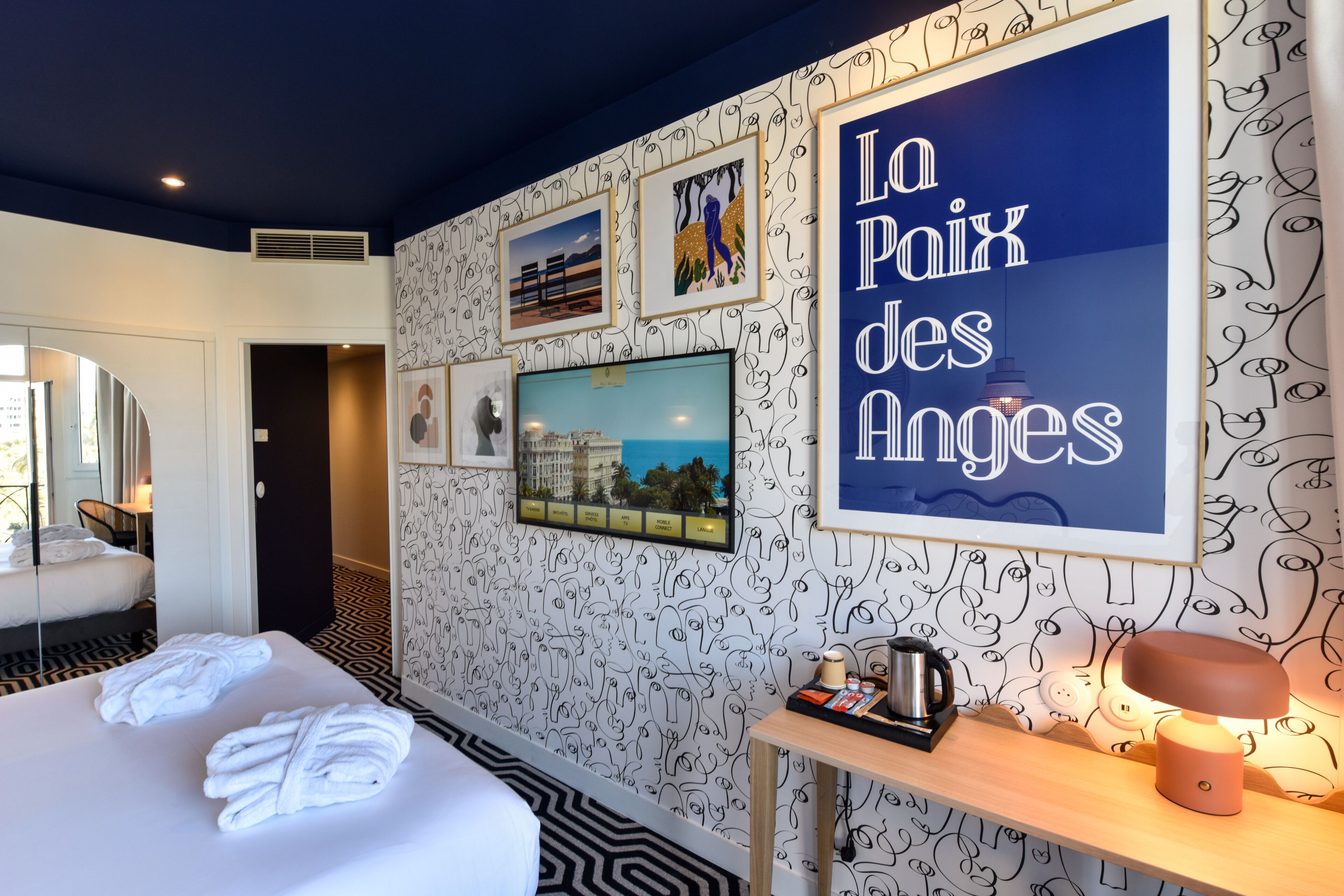 A hotel room with a bed, a desk with a lamp, a television, and artwork on the wall.